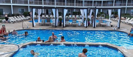 Une piscine entourée de chaises longues et de parasols, elle-même bordée d’un immeuble doté de nombreux balcons.