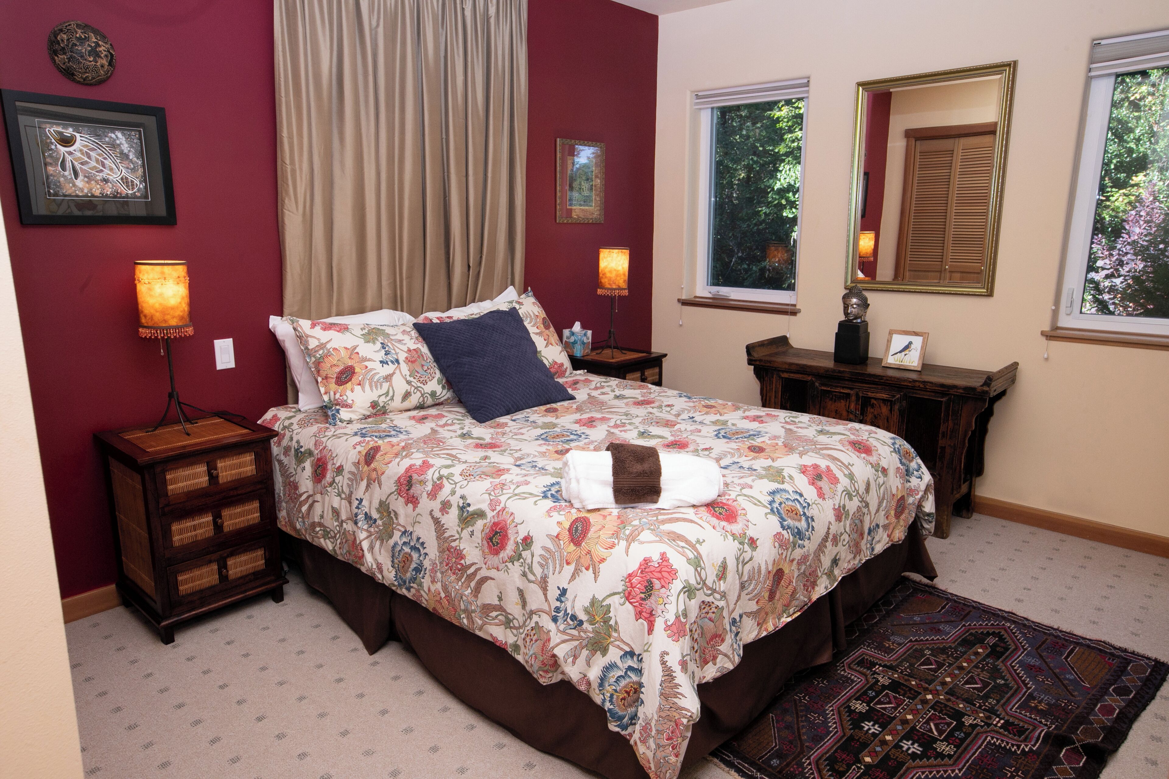 Une chambre à coucher avec une couvre-lit à motifs floraux, une lampe de chevet, une table de chevet en bois, une commode et une fenêtre donnant sur des arbres.
