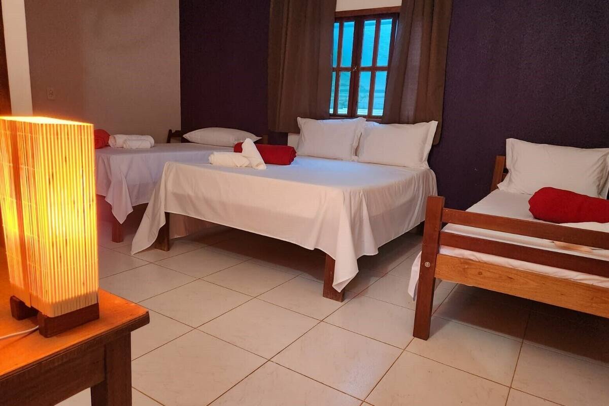 Quarto de hotel com duas camas de solteiro, um banco de madeira, uma mesa com abajur e uma janela com cortinas.