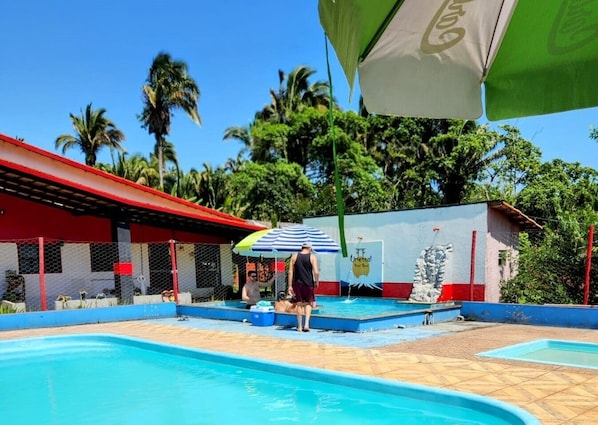 A swimming pool area with a covered seating area, a mural, and a person under a striped umbrella.