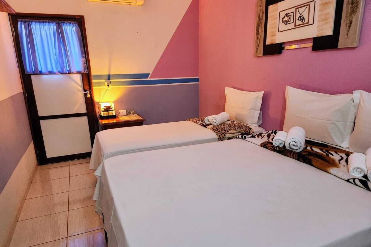 Quarto de hotel com duas camas, mesa de cabeceira com abajur, porta com cortinas e parede com quadros.