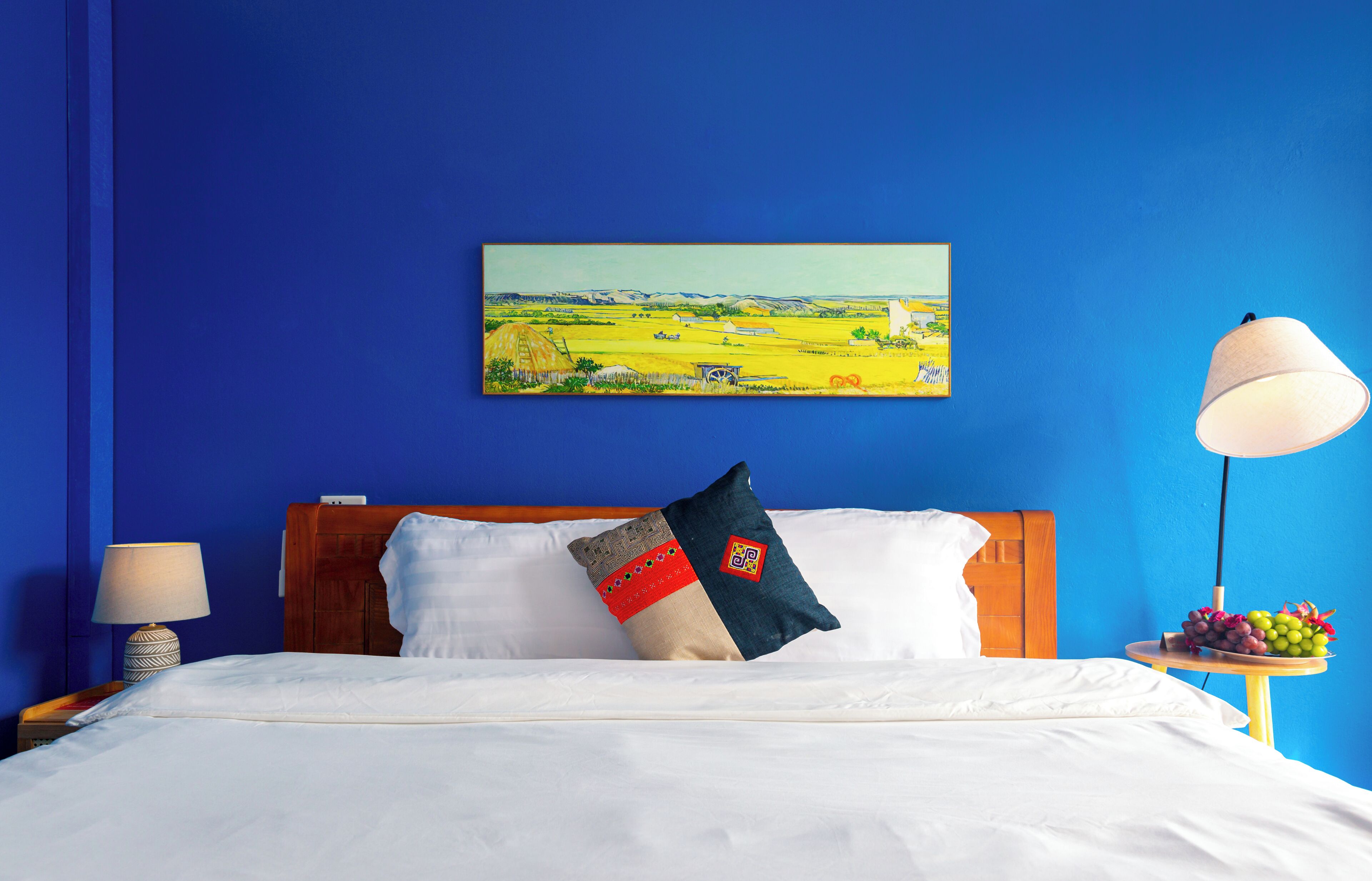 Una habitación de hotel con una cama, una mesita de noche con lámpara, una mesa con fruta y un cuadro en la pared.