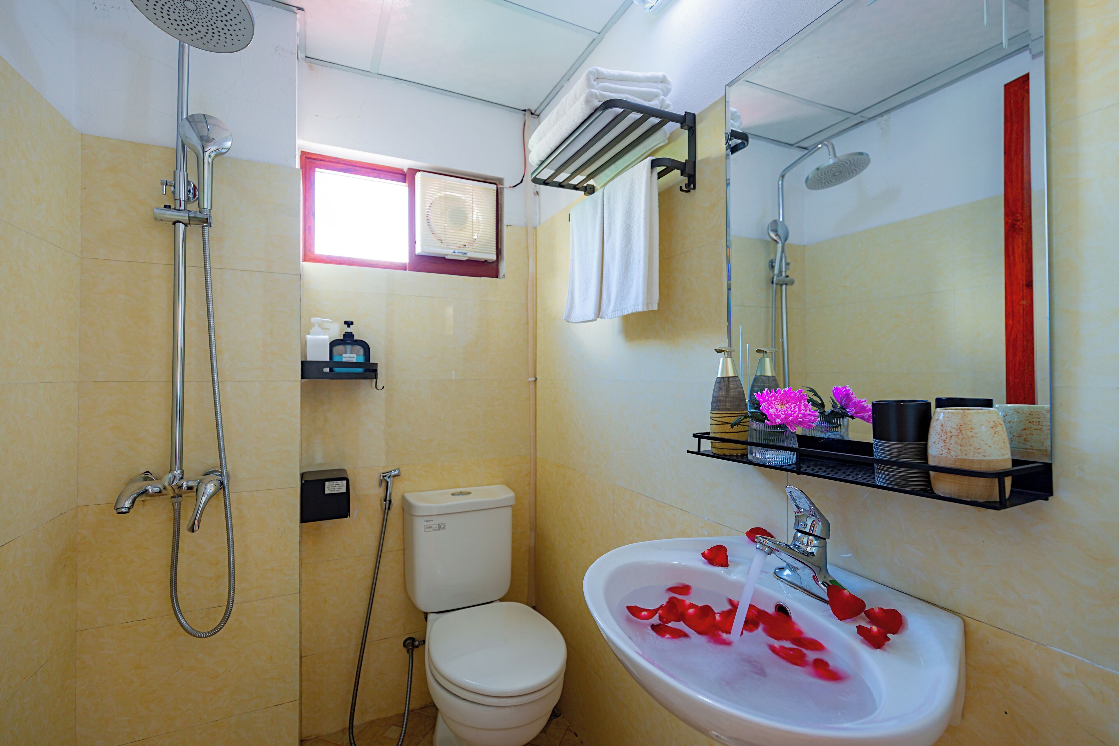 Un baño con ducha, inodoro, lavamanos con pétalos de rosa y un espejo.
