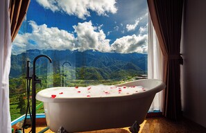 A bathtub with rose petals, a showerhead, and a view of mountains through a window. - Moc Home Sapa Center (Sa Pa)