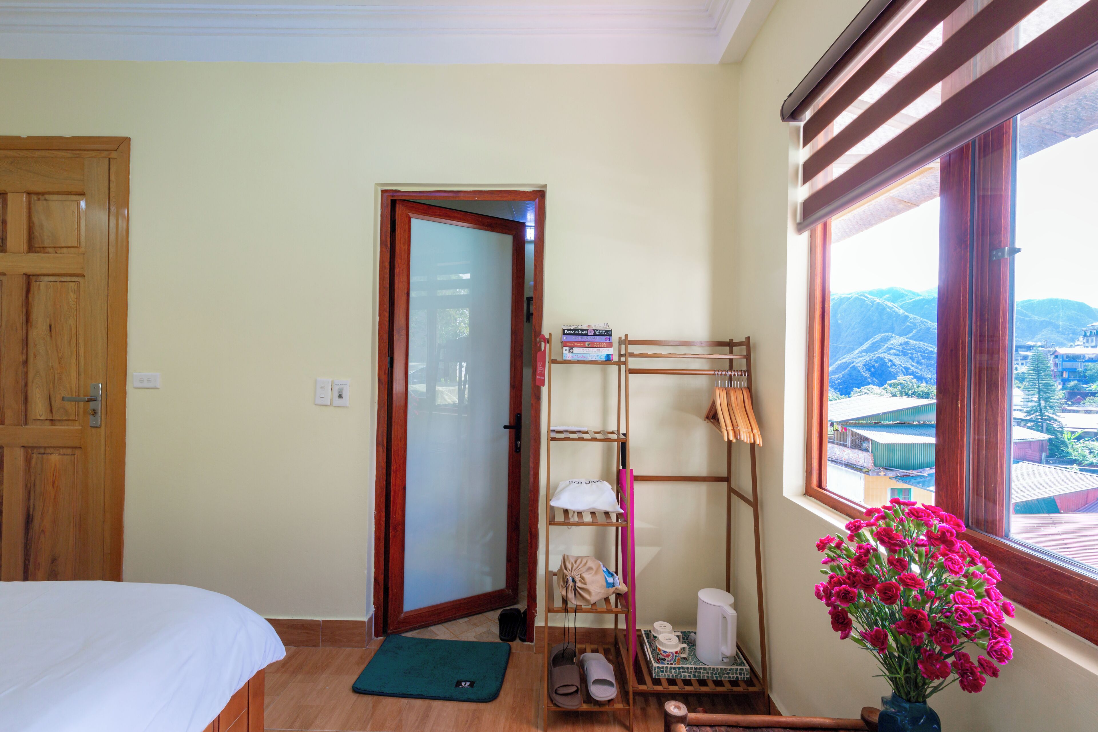 A hotel room with a bed, a wooden door, a shelf with towels and toiletries, a window with a view of buildings, and a vase with flowers.