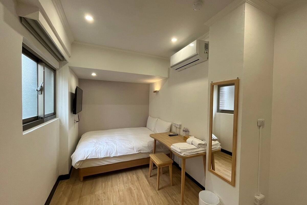 A small hotel room with a bed, a bedside table with a lamp, a mirror, and a window.
