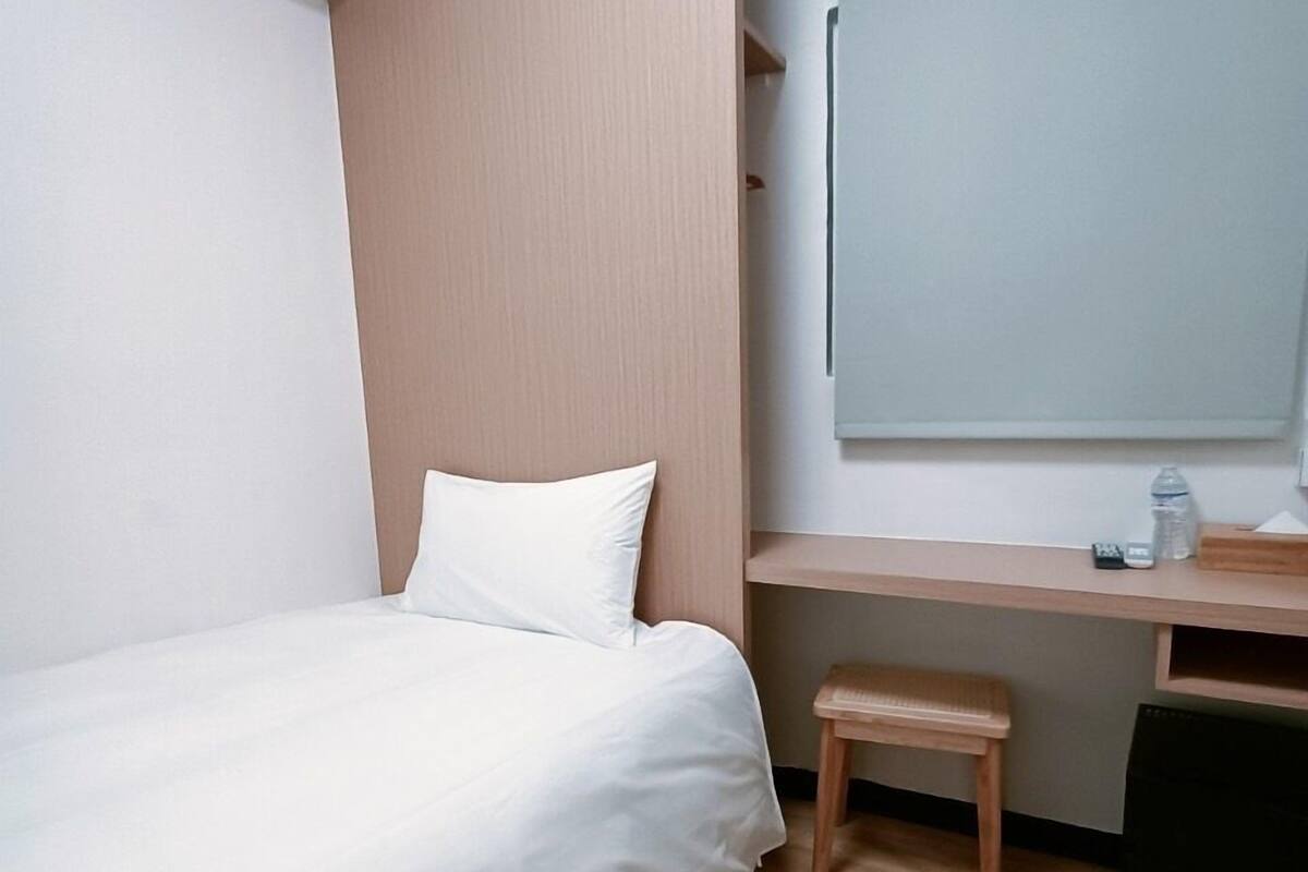 A small, neatly arranged bedroom with a single bed, a wooden desk, and a small stool.