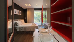 Une chambre d’hôtel moderne, équipée d’un lit, d’une table à manger dressée avec des assiettes et des verres, et d’une grande fenêtre donnant sur un paysage verdoyant.