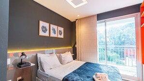 Une chambre d’hôtel moderne avec un grand lit, une table de chevet avec une lampe, des œuvres d’art encadrées au mur et un balcon avec vue.