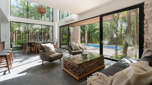 A spacious living area with a glass coffee table, a sofa, and a dining table with chairs. Large windows offer views of a swimming pool and trees. - Twelve O’Clock Luxury Villas (Tulum)