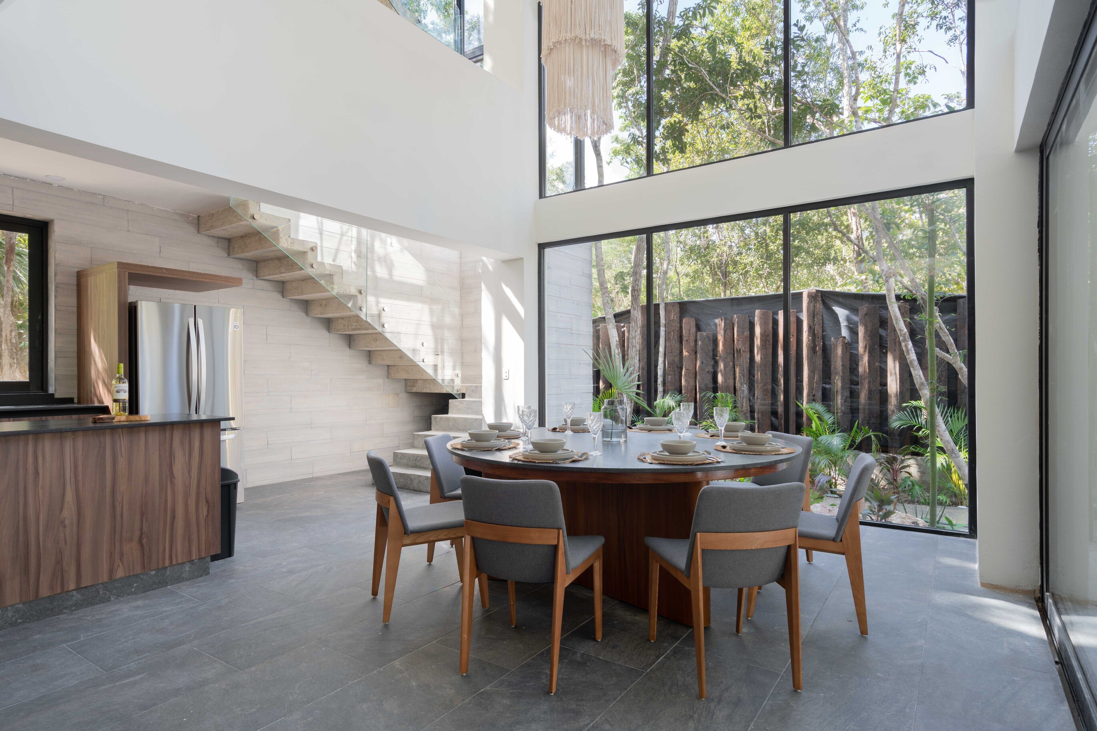 Une cuisine moderne dotée d’un îlot central, d’un coin repas avec une table ronde et des chaises, et d’une grande fenêtre donnant sur un jardin.