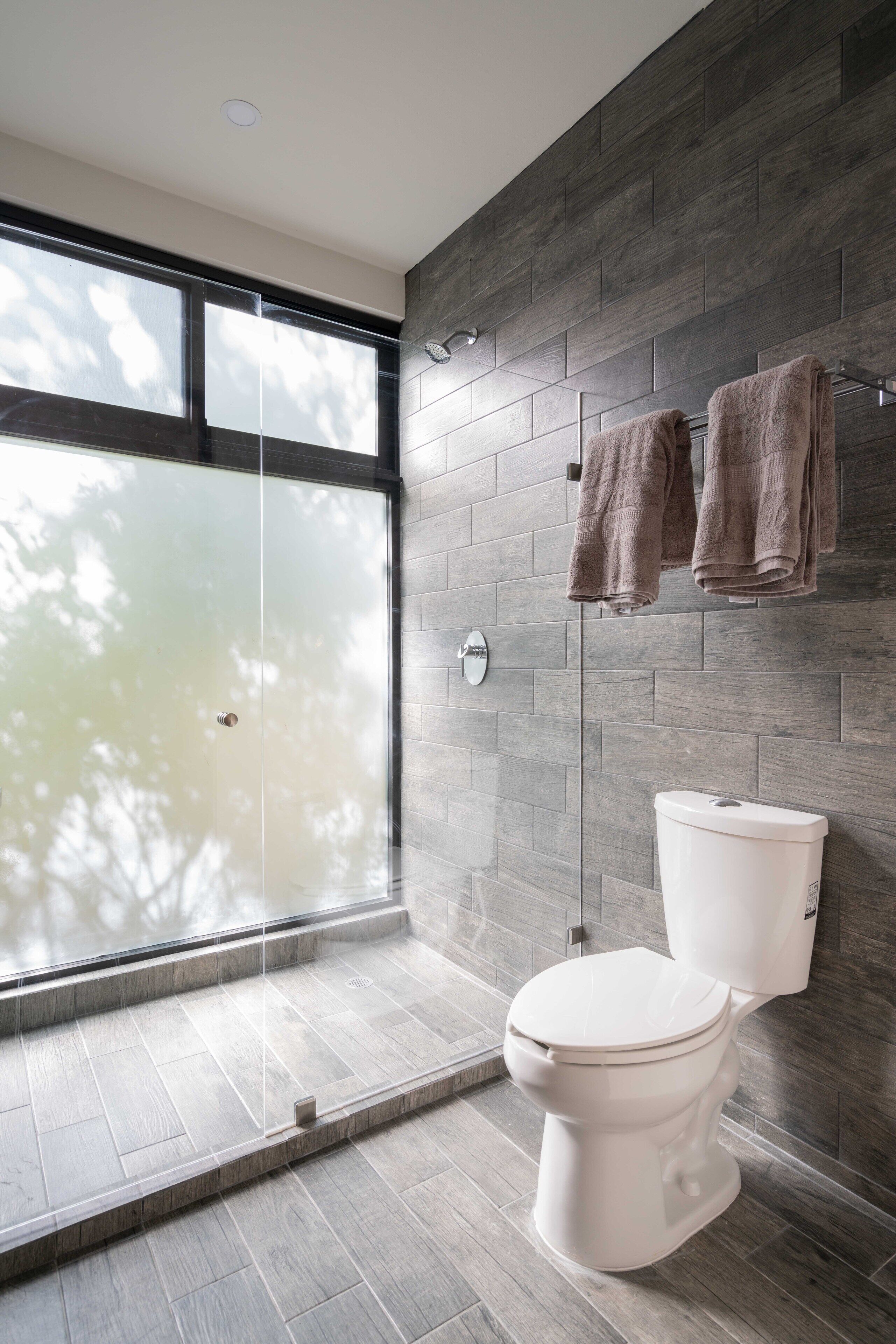 A modern bathroom with a large window, a glass shower enclosure, and a toilet against a tiled wall.