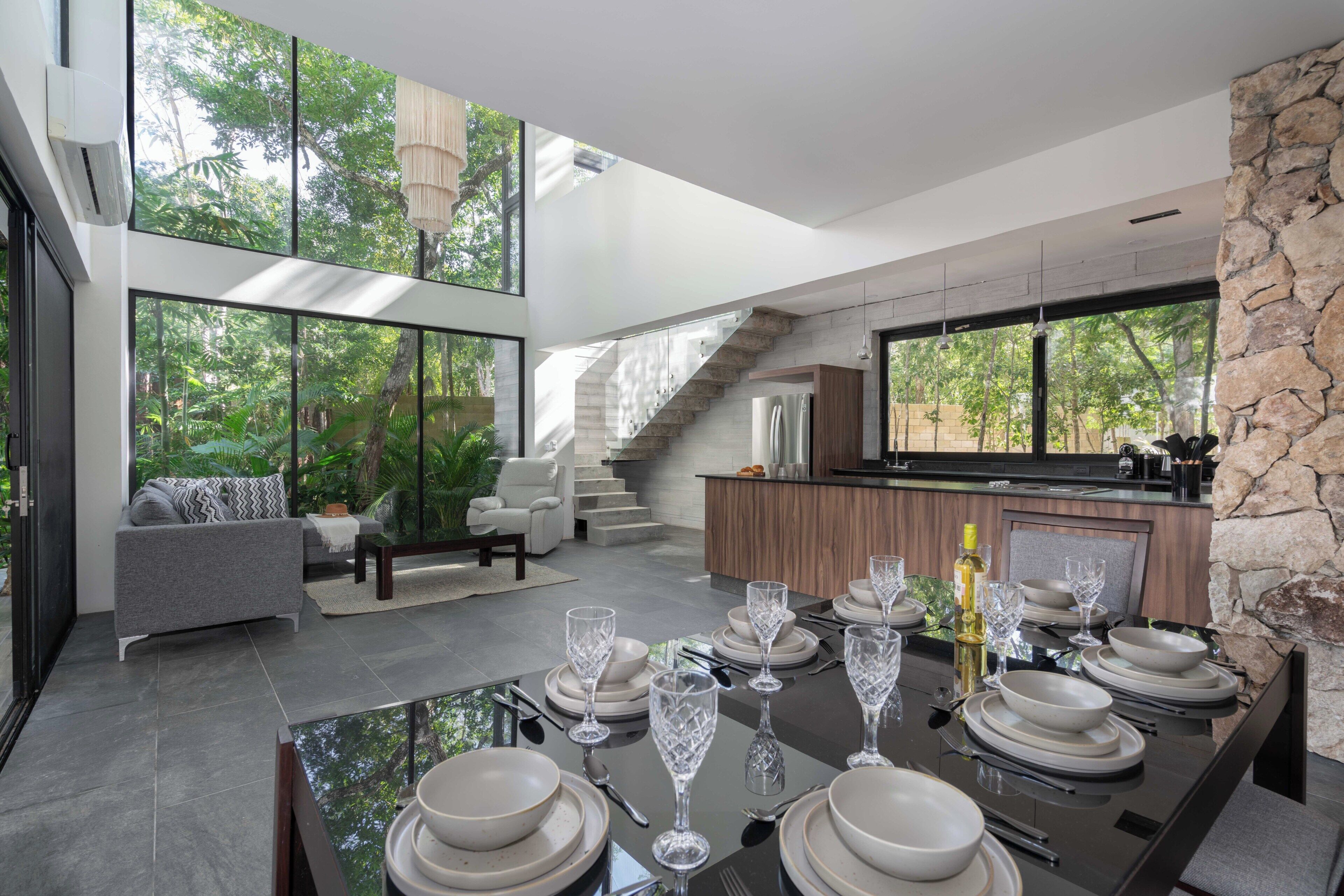 Un espace de repas moderne avec une table en verre dressée pour un repas, une cuisine avec un mur d’accent en pierre et un escalier menant à un niveau supérieur.