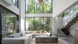 A modern living room with a sectional sofa, a marble coffee table, and large windows offering a view of the outdoors. - Twelve O’Clock Luxury Villas (Tulum)