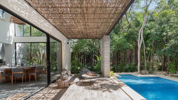 A modern outdoor pool area with a wooden deck, lounge seating, and a glass-enclosed dining area. - Twelve O’Clock Luxury Villas (Tulum)
