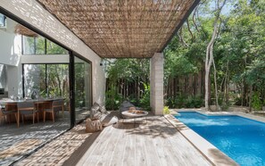 A modern outdoor pool area with a wooden deck, lounge seating, and a glass-enclosed dining area. - Twelve O’Clock Luxury Villas (Tulum)