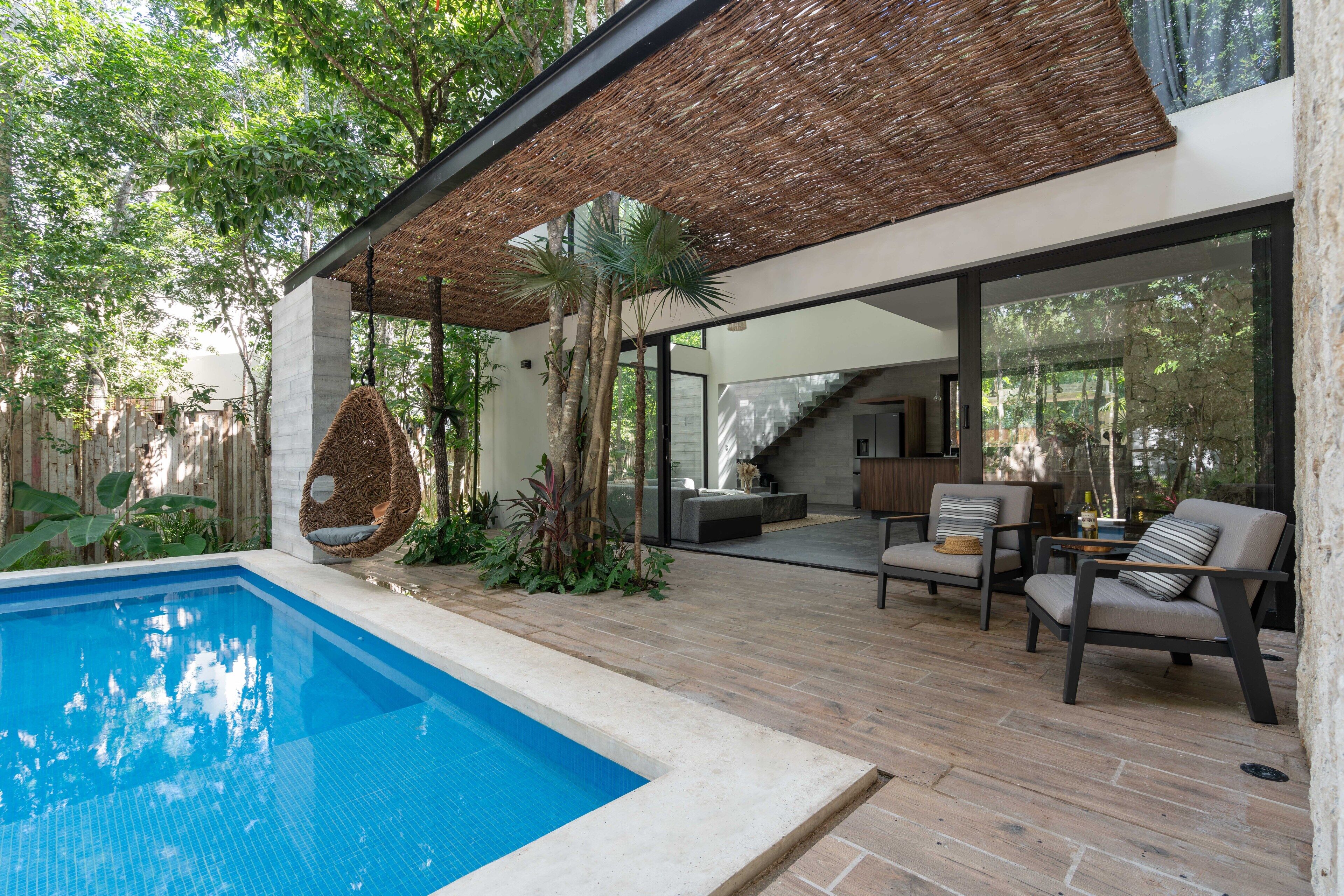 A modern outdoor pool area with a clear blue pool, wooden decking, and a covered seating area with chairs and a hanging chair.