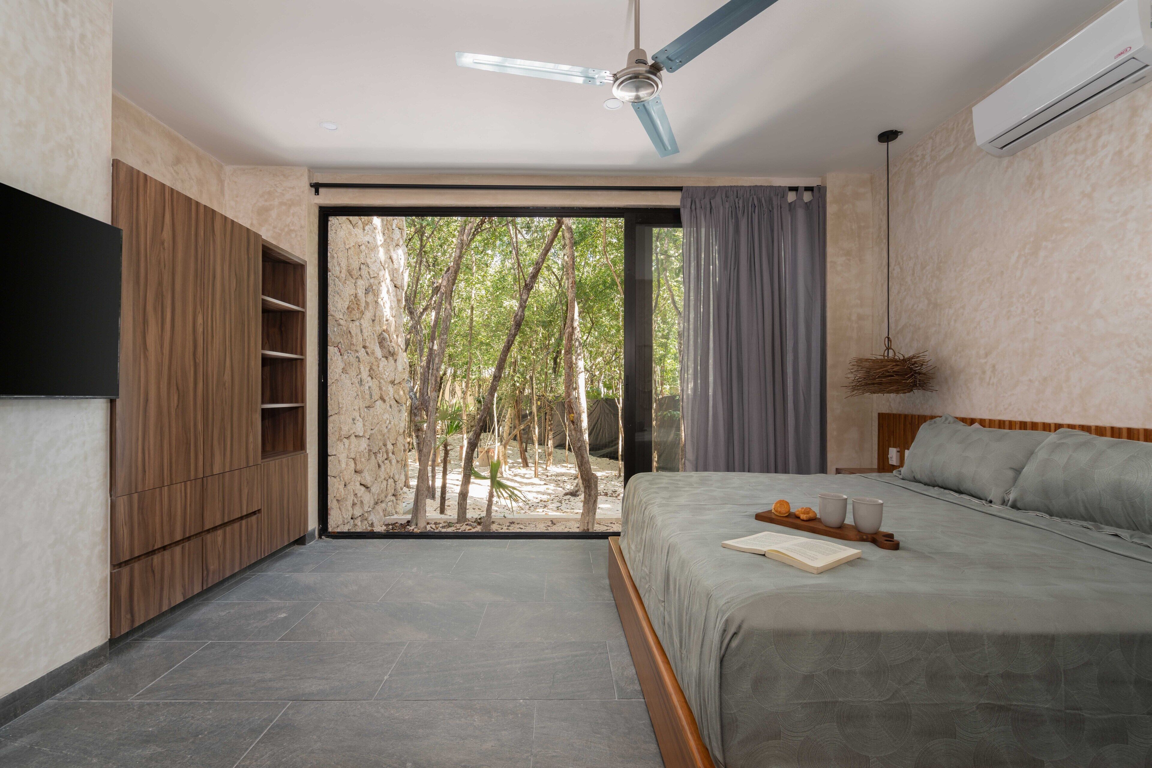Un dormitorio con cama, ventilador de techo, televisor y vistas a árboles a través de un ventanal.