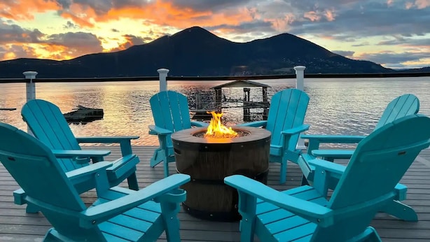 A deck with four turquoise Adirondack chairs and a central fire pit, overlooking a calm lake and a mountain at sunset. - Smiling Otter Retreat! Large Lakefront Luxury House.Pool.Spa.Sauna.Games..Pets (Clearlake)