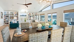 A spacious living room with a large wooden dining table, wicker chairs, and a chandelier. There are bookshelves filled with books and decorative items, and a view of the outdoors through large windows. - Smiling Otter Retreat! Large Lakefront Luxury House.Pool.Spa.Sauna.Games..Pets (Clearlake)