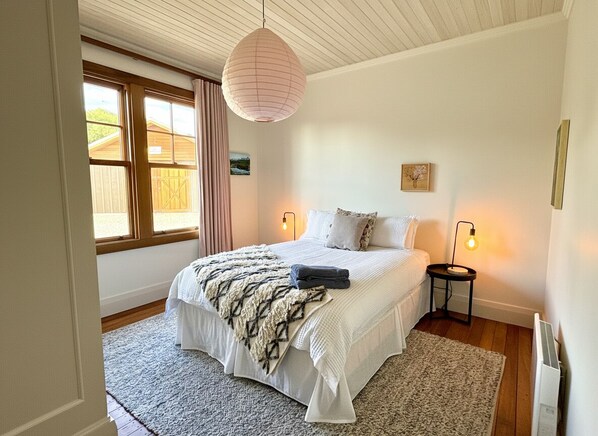 A bedroom with a large bed, a window with curtains, a hanging light fixture, and a bedside table with a lamp.