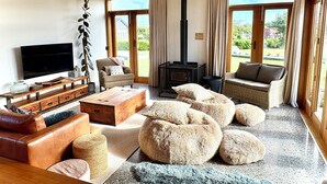 Ein geräumiges Wohnzimmer mit großem Fernseher, bequemen Sitzmöglichkeiten, einem Kamin und großen Fenstern mit Blick ins Freie.