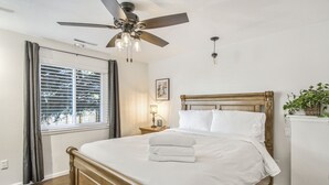 A bedroom with a wooden bed, a ceiling fan, a window with curtains, a nightstand with a lamp, and a small plant on a cabinet. - Mid-Farmers Park (Colorado Springs)