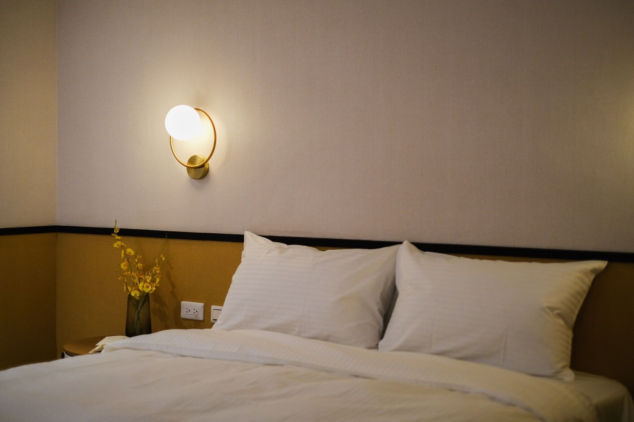 Habitació d'hotel amb llit, roba de llit blanca, llum penjada a la paret i banyador de flors a la nit.