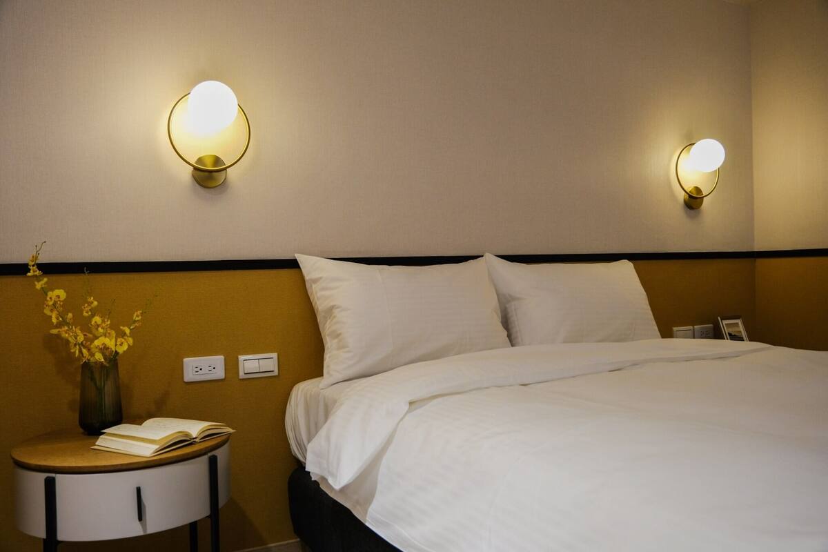 A hotel room with a neatly made bed, two wall-mounted reading lamps, a small round table with a book and a vase of flowers, and a power outlet on the wall.