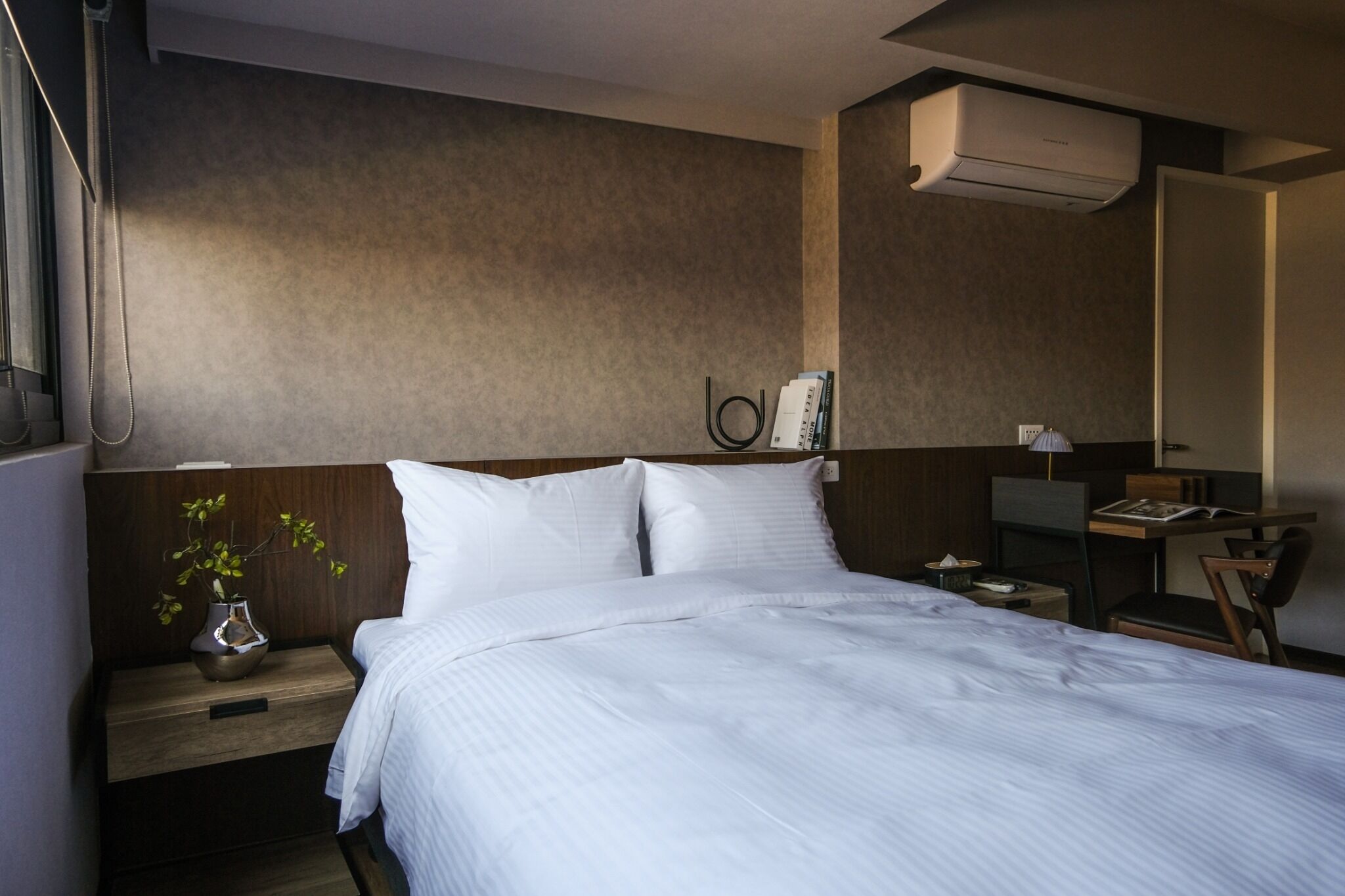 A hotel room with a large bed, a desk, a chair, a nightstand with a vase, and an air conditioning unit.