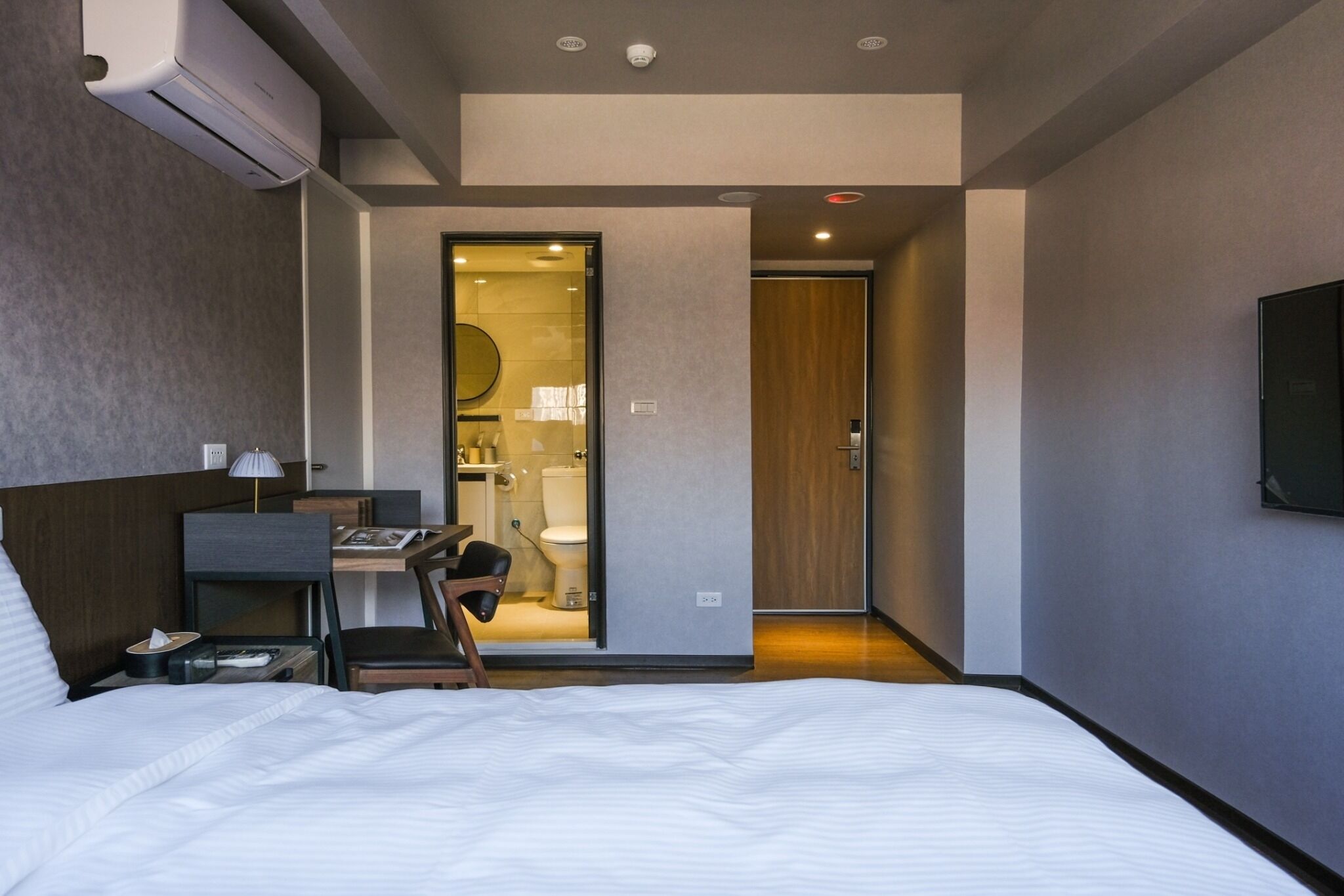 A modern hotel room with a large bed, a desk, a chair, and a bathroom visible through an open door.