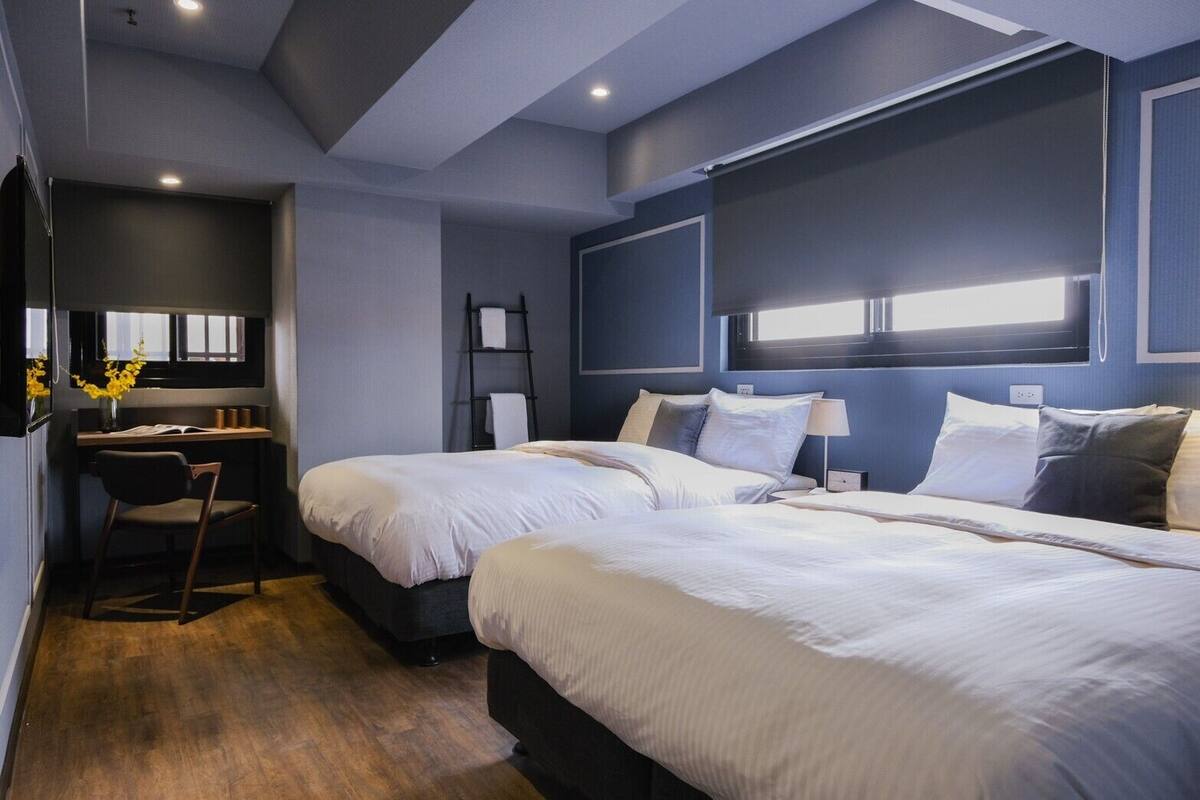 A hotel room with two beds, a desk, a chair, and a window.