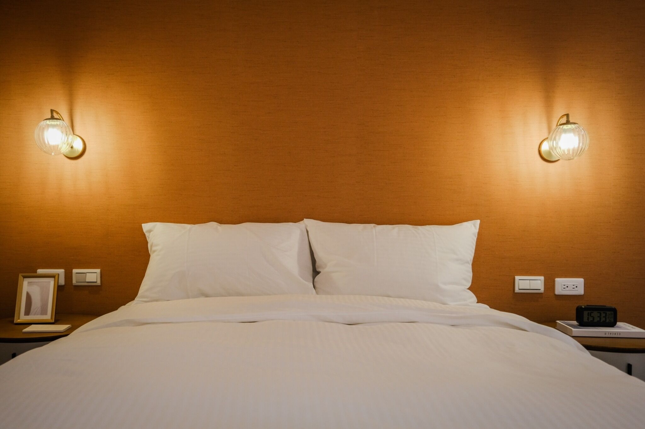 A neatly made bed with white linens, two wall-mounted light fixtures, a bedside table with a framed picture, a nightstand with a clock, and a power outlet.