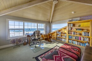 A room with a large window overlooking the ocean, a wooden ceiling, a bookshelf, and a cozy seating area.