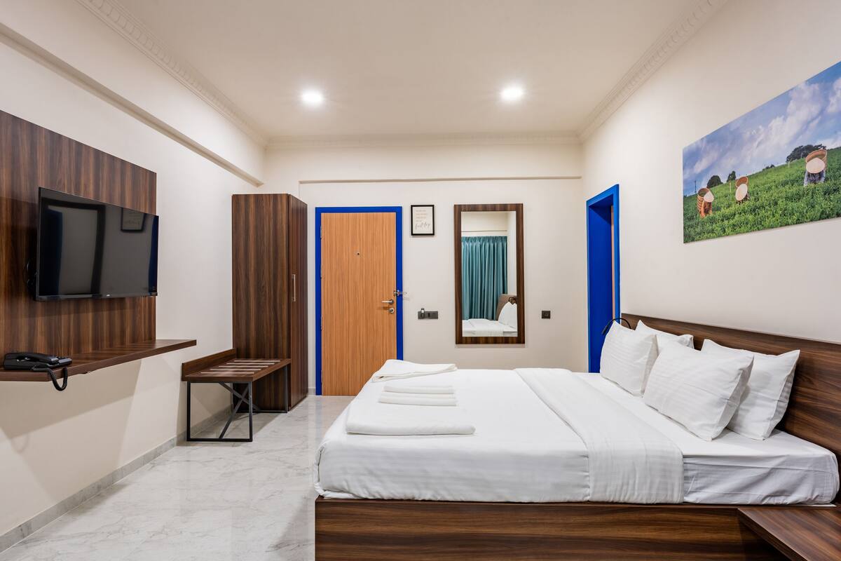 A hotel room with a bed, a desk with a phone, a TV mounted on the wall, a wardrobe, and a painting on the wall.
