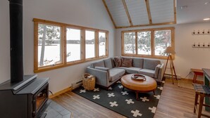 A cozy living room with a fireplace, large windows, a sectional sofa, and a wooden floor.