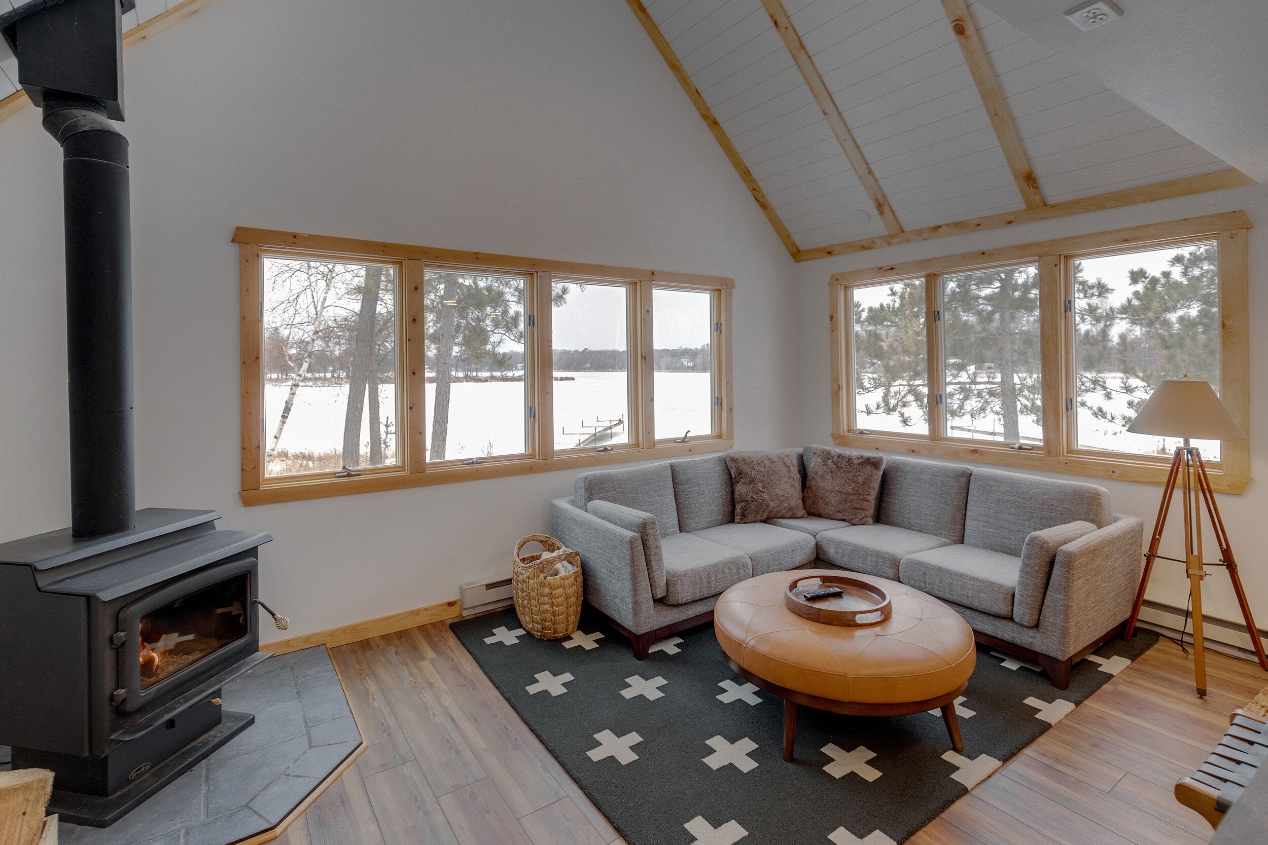 A cozy living room with a fireplace, a gray sofa, a wooden floor, and large windows overlooking a snowy landscape.