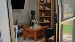 A living room with a wooden coffee table, a bookshelf, a television mounted on the wall, and a sliding glass door leading outside.