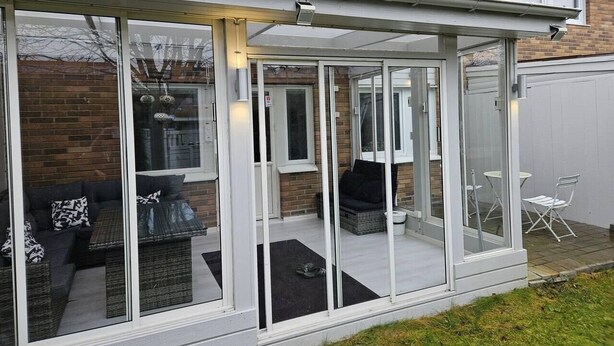 A covered patio area with a glass enclosure, outdoor seating, and a brick wall in the background.