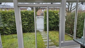 A conservatory with sliding glass doors, a stone pathway, and a view of a hedge and gate.