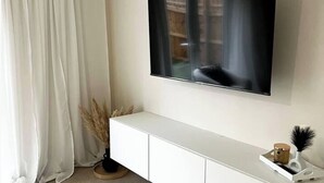 Moderni olohuone, jossa seinälle on asennettu taulutelevisio, huoneessa on valkoinen konsolipöytä ja ikkunassa läpikuultavat verhot.