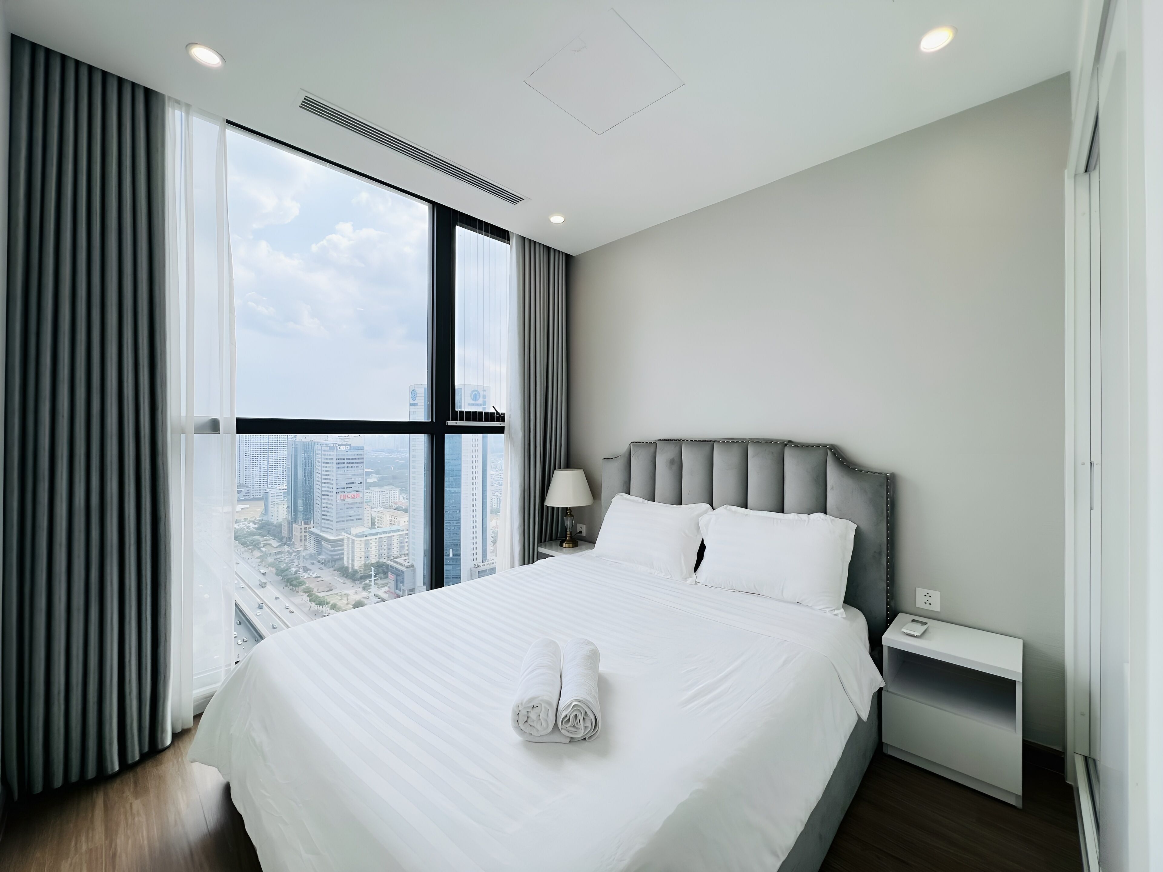 A modern hotel room with a large bed, a nightstand, a lamp, and a view of the city through large windows.
