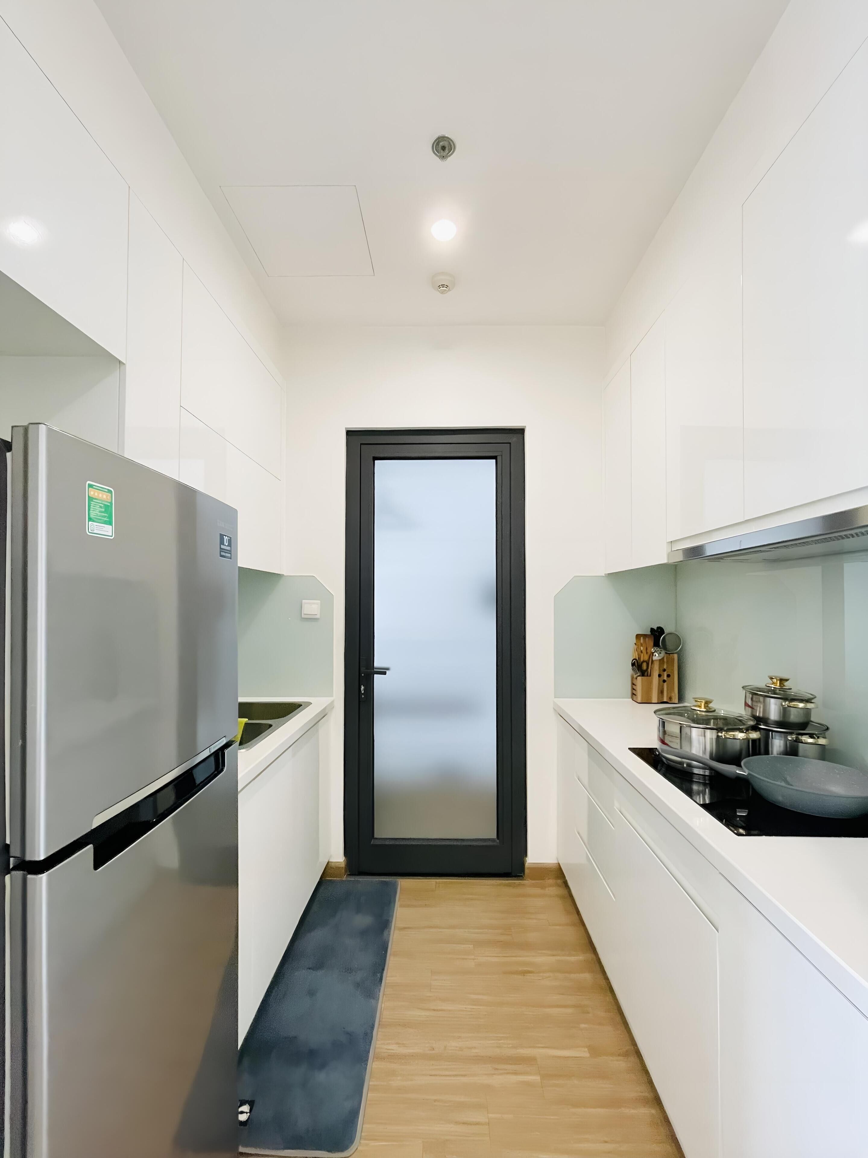 Una cocina estrecha con gabinetes blancos, un refrigerador de acero inoxidable, una puerta negra y piso de madera.