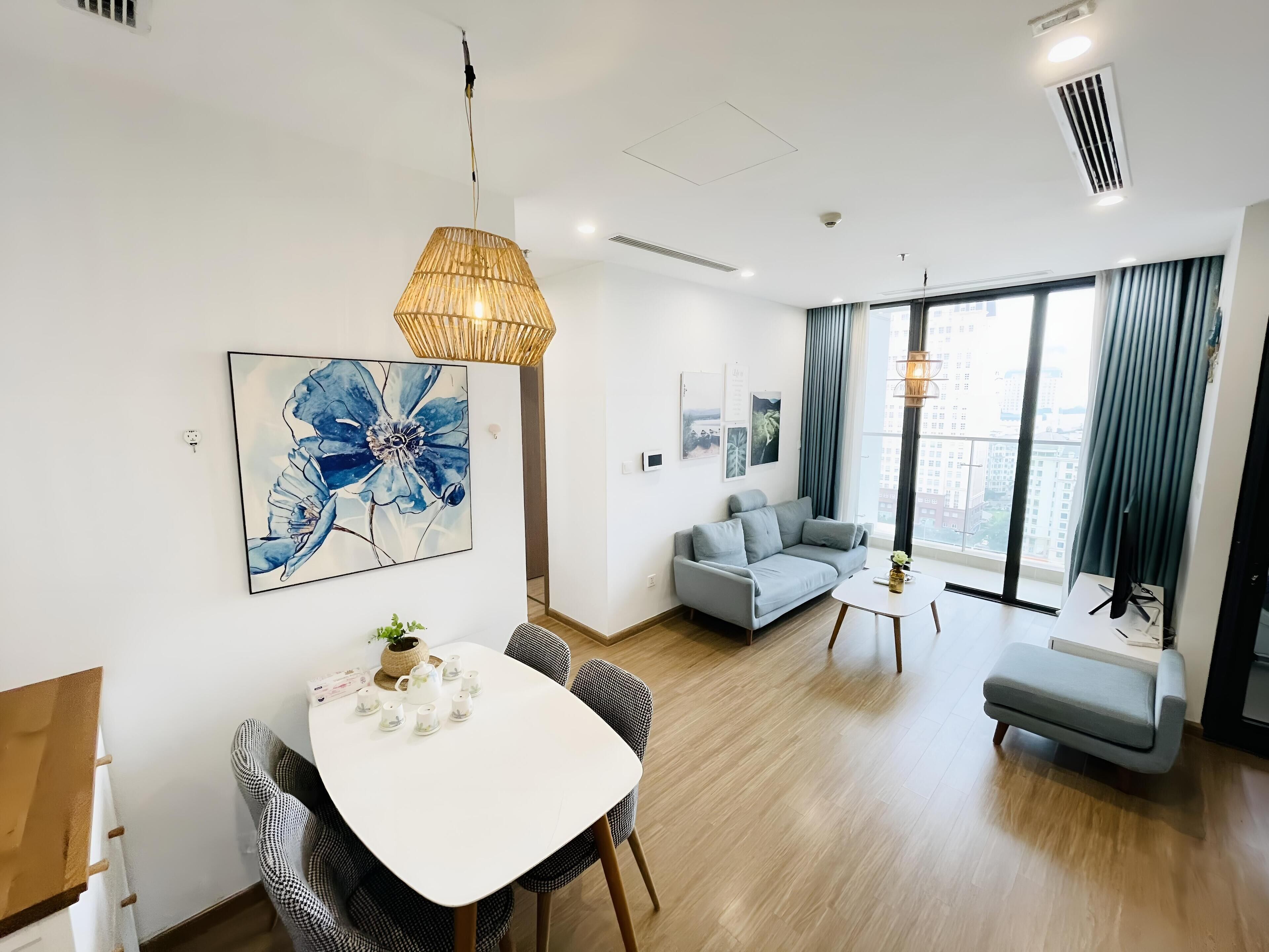 A modern living room with a white sofa, a dining table with chairs, a large window with curtains, and a city view.