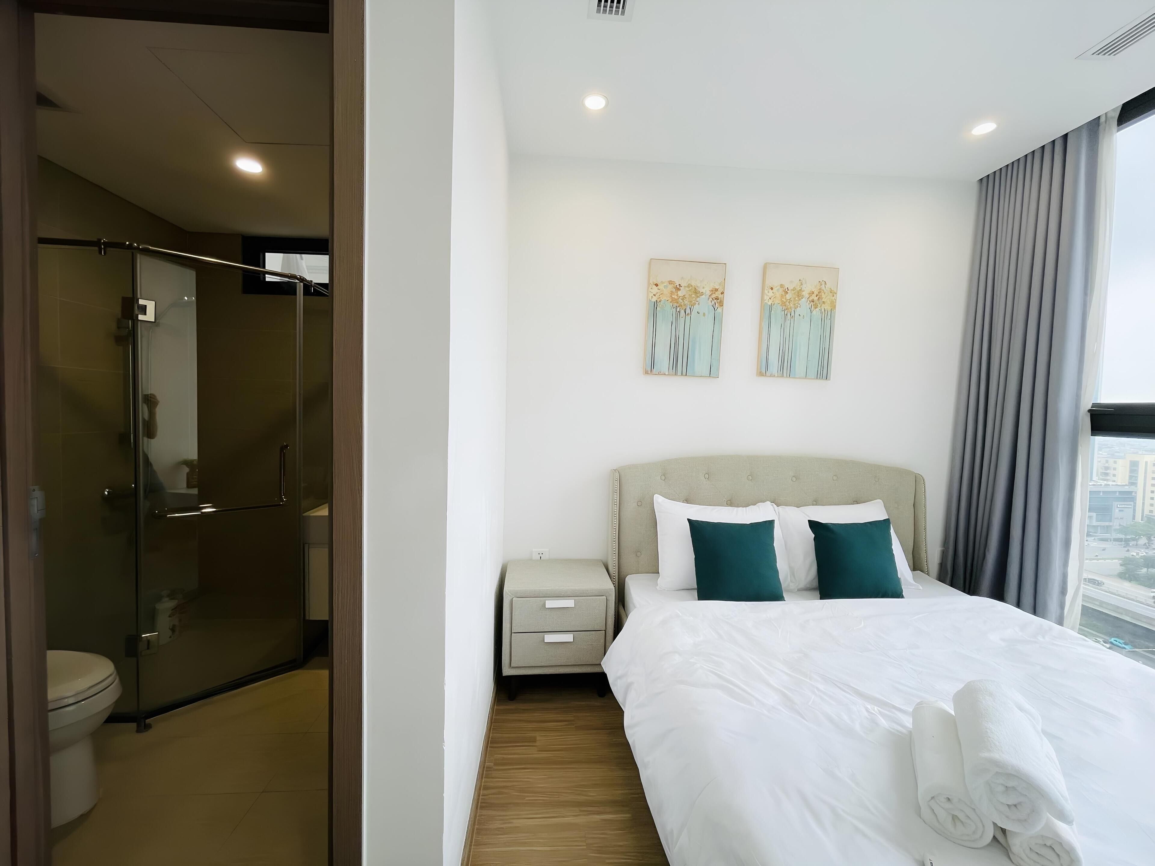 Habitación de hotel con cama, mesita de noche, ducha con mampara de vidrio y dos cuadros colgados en la pared.