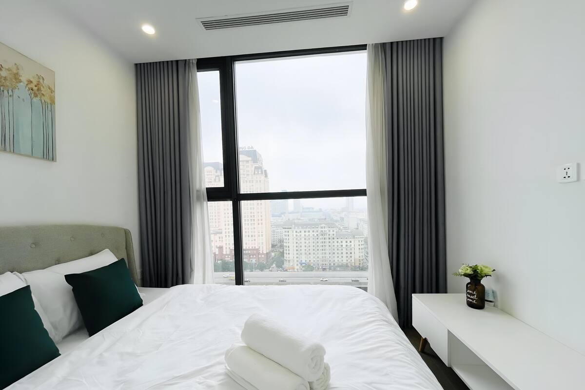 Ein Hotelzimmer mit einem großen Bett, einem Nachttisch, einem Fenster mit Vorhängen und Blick auf die Stadt.