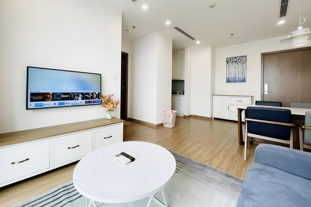 Ein modernes Wohnzimmer mit Flachbildfernseher, einem weißen Couchtisch, einer Couch, einem Essbereich mit Tisch und Stühlen und einer Küche im Hintergrund.