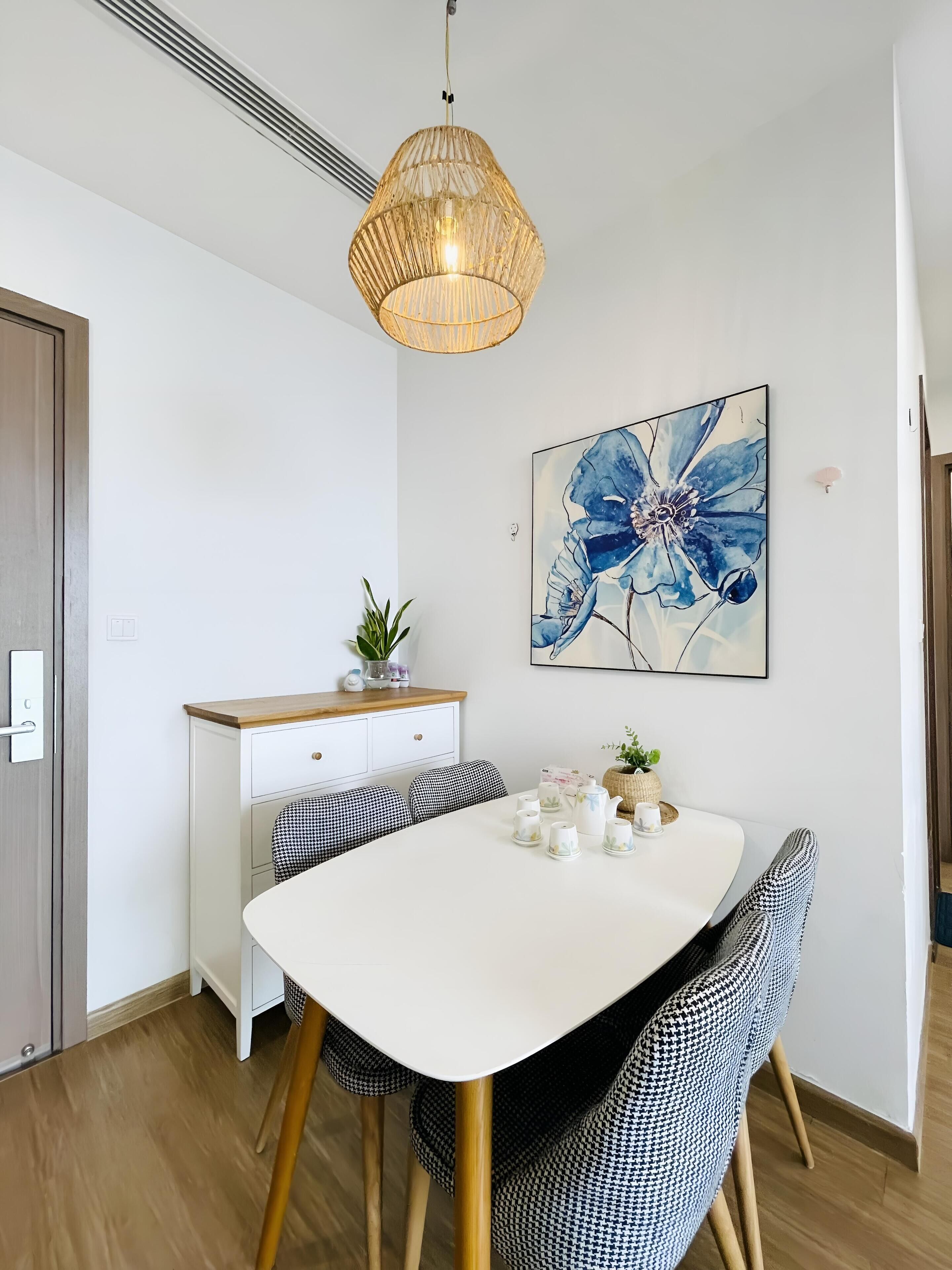 Un espace repas moderne avec une table blanche, des chaises à motifs noir et blanc, un luminaire suspendu et un tableau floral bleu accroché au mur.