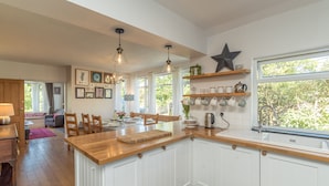 Una cocina moderna con armarios blancos, encimeras de madera y un ventanal grande. Hay lámparas colgantes, una decoración con forma de estrella y varios utensilios de cocina en estantes.