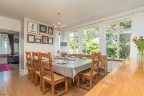 A dining area with a wooden table and chairs, framed artwork on the walls, and large windows with a view of greenery outside. - Elegant Holiday Cottage on the Banks of the River Artro | Artro (Llanbedr)